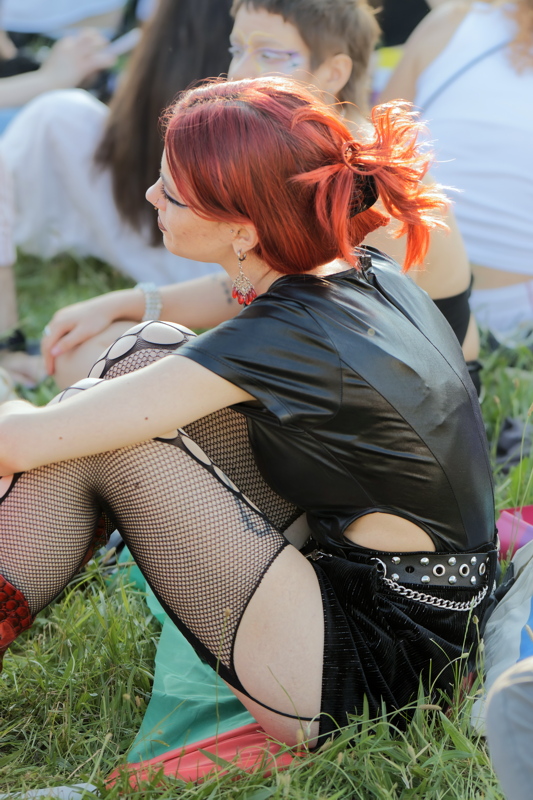 23 - Person with vibrant red hair wearing a black sleeveless top, a studded belt, and fishnet tights, attendee at a Pride parade
